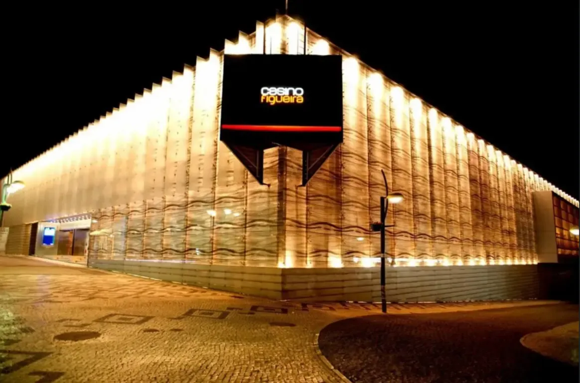 Edifício de painéis ondulados iluminados de baixo à noite, com o letreiro casino figueira na esquina e calçada desenhada à frente.