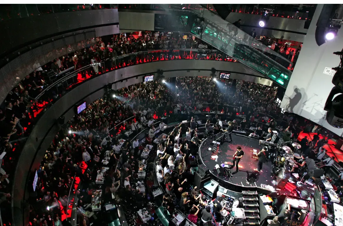 Sala circular vista de cima, com público em vários pisos à volta de um palco redondo onde toca uma banda, sob focos de luz.