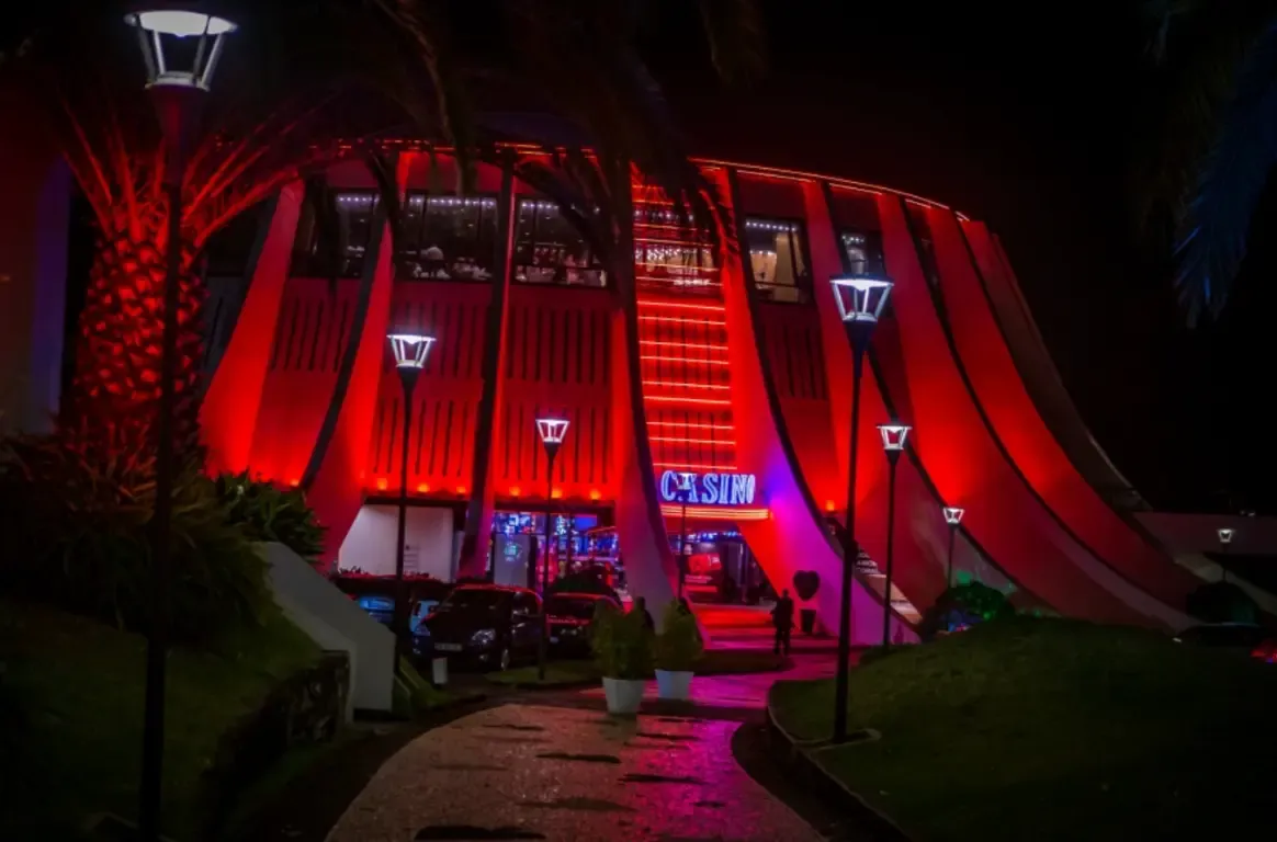 Fachada curva iluminada a vermelho à noite, com o letreiro CASINO em azul, candeeiros ao longo de um caminho e palmeiras à frente.