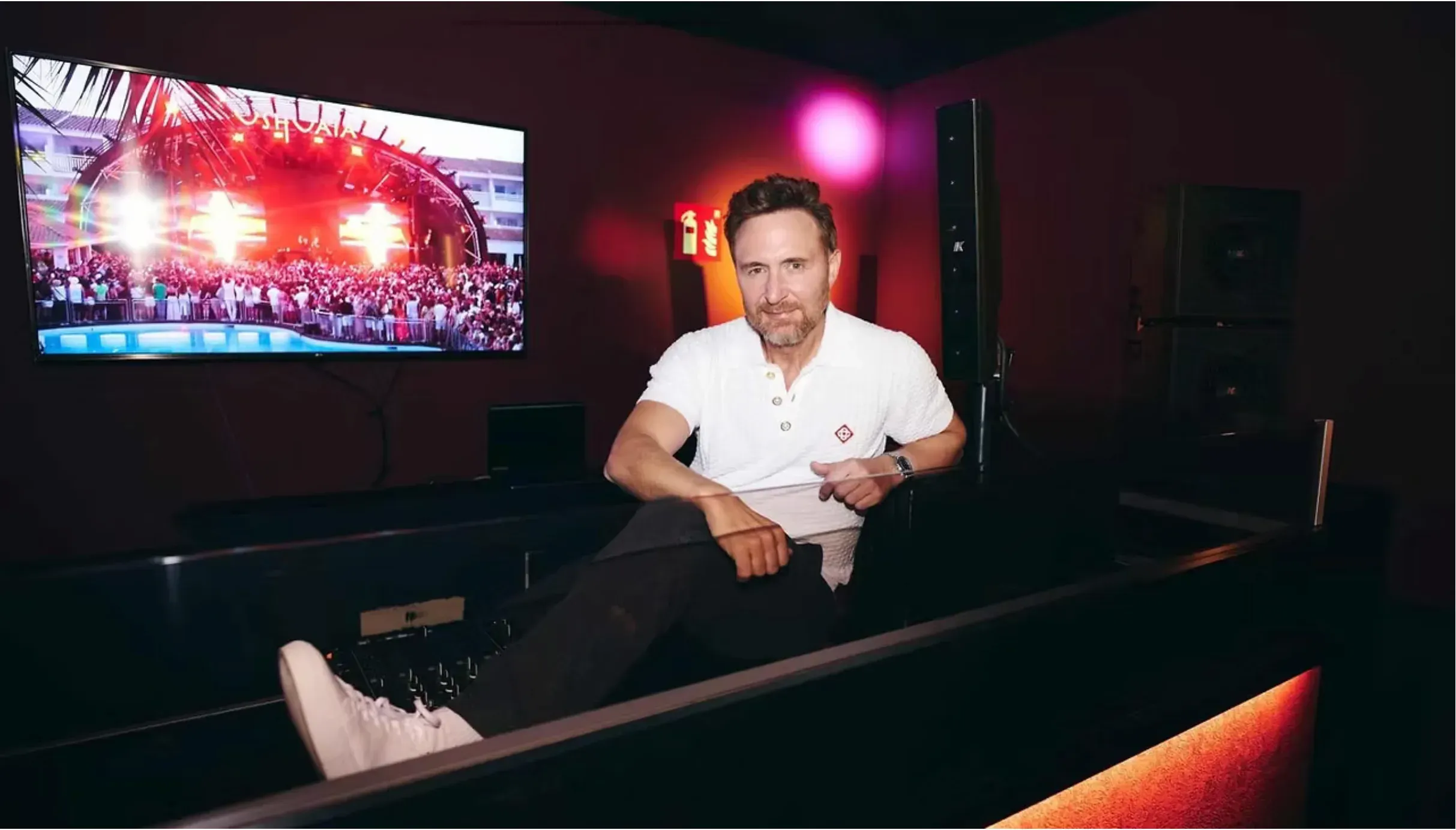 David Guetta sentado numa cabine de DJ escura, ao lado de colunas com o símbolo da K-array e de um ecrã que mostra um concerto ao ar livre, junto a uma piscina.