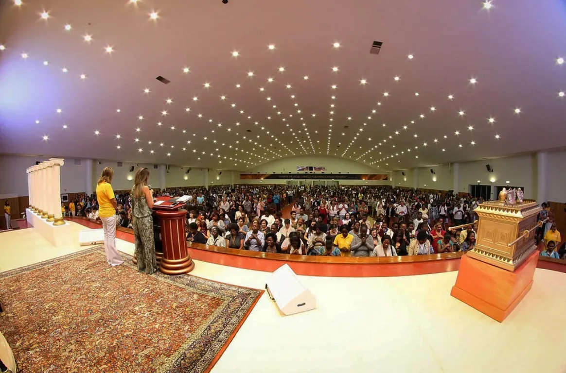 Salão de culto visto do palco, com duas pessoas junto ao púlpito, uma peça dourada à direita e a assembleia de pé sob um teto de pontos de luz.