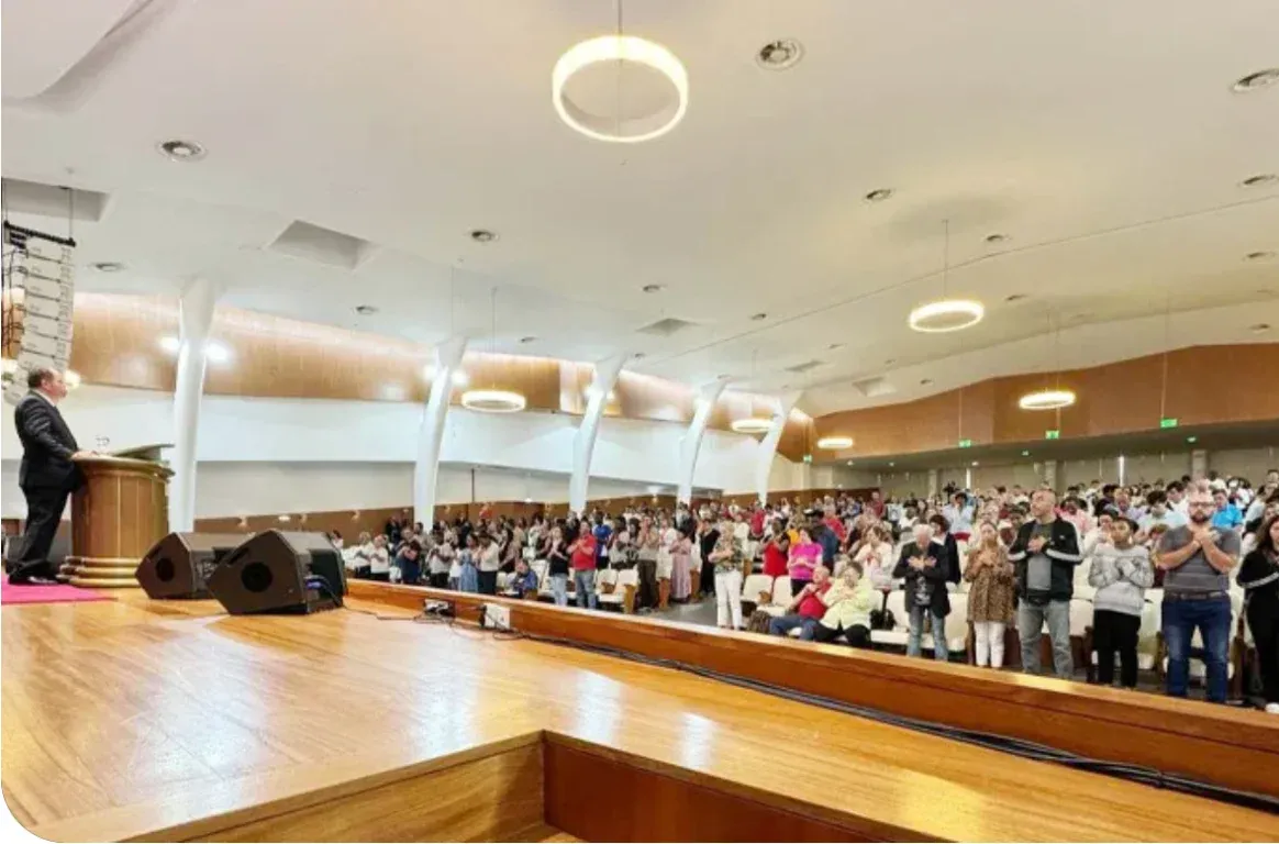 Salão com o orador no púlpito, colunas de som no palco de madeira e a assembleia de pé entre pilares brancos e candeeiros circulares.