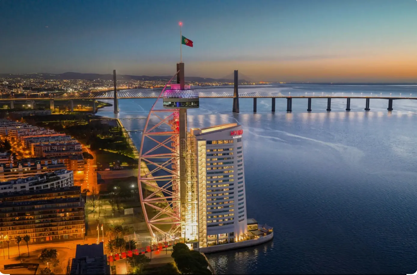 A Torre Vasco da Gama e o edifício do hotel ao anoitecer, vistos do alto, com o rio e a Ponte Vasco da Gama iluminada ao fundo.