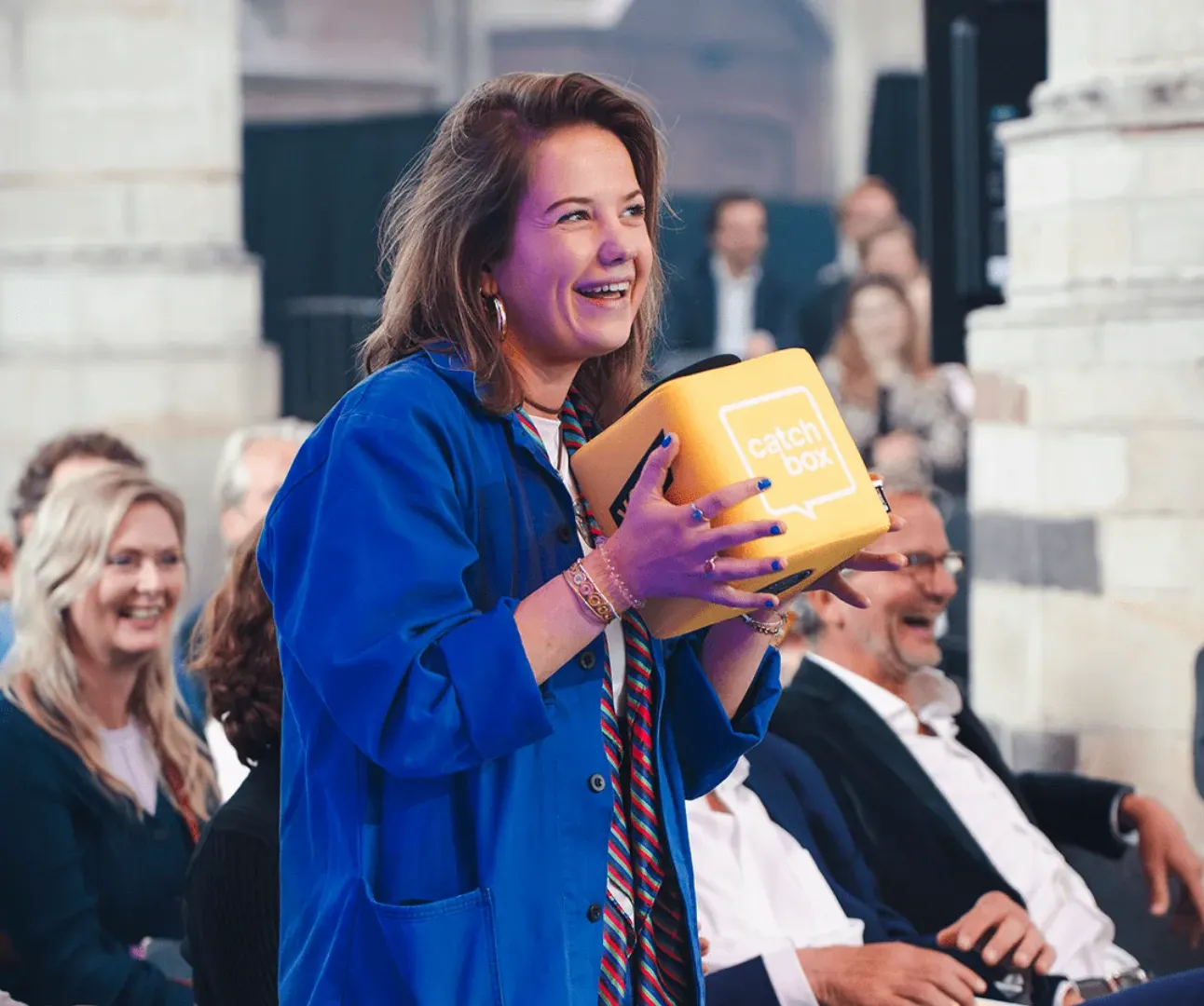 Mulher de casaco azul a sorrir com um cubo de microfone amarelo nas mãos, no meio do público de um evento.