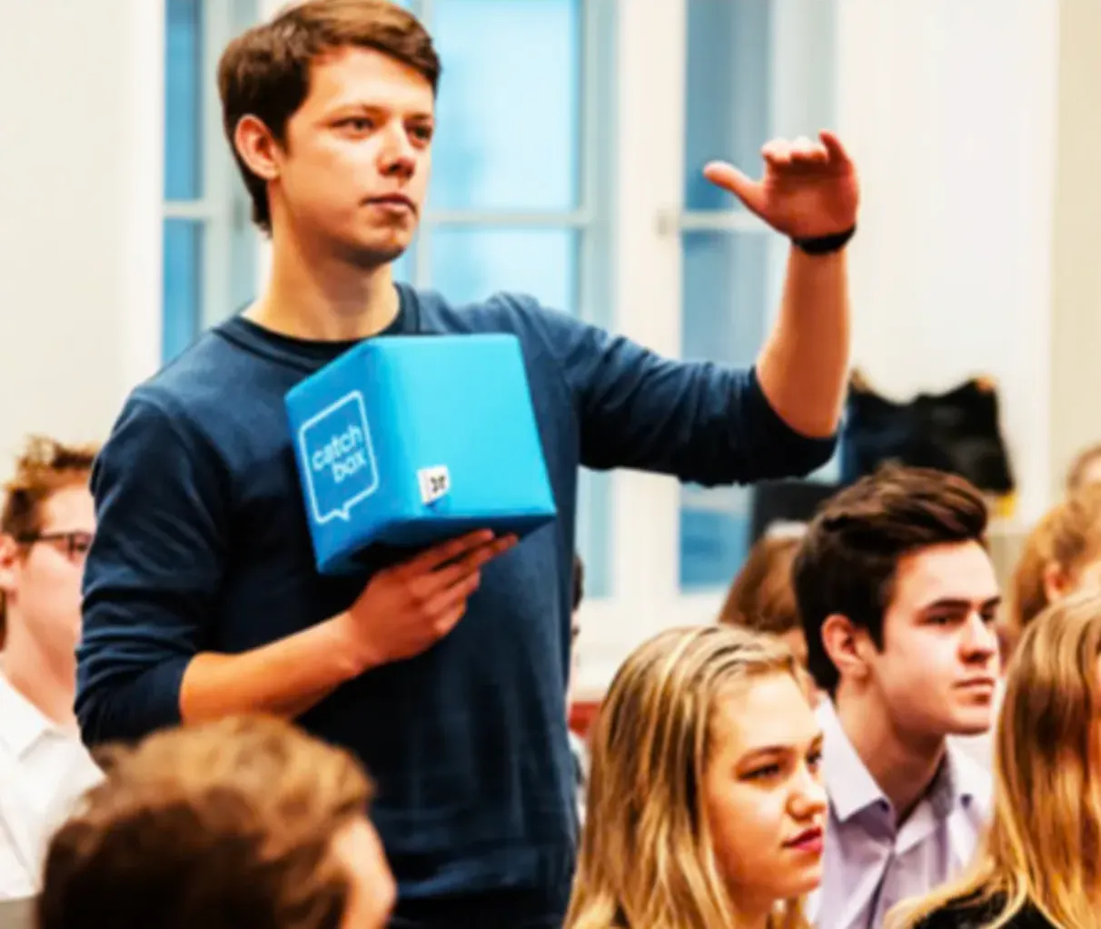 Jovem de pé no meio de um grupo de estudantes, a falar com um cubo de microfone azul na mão.