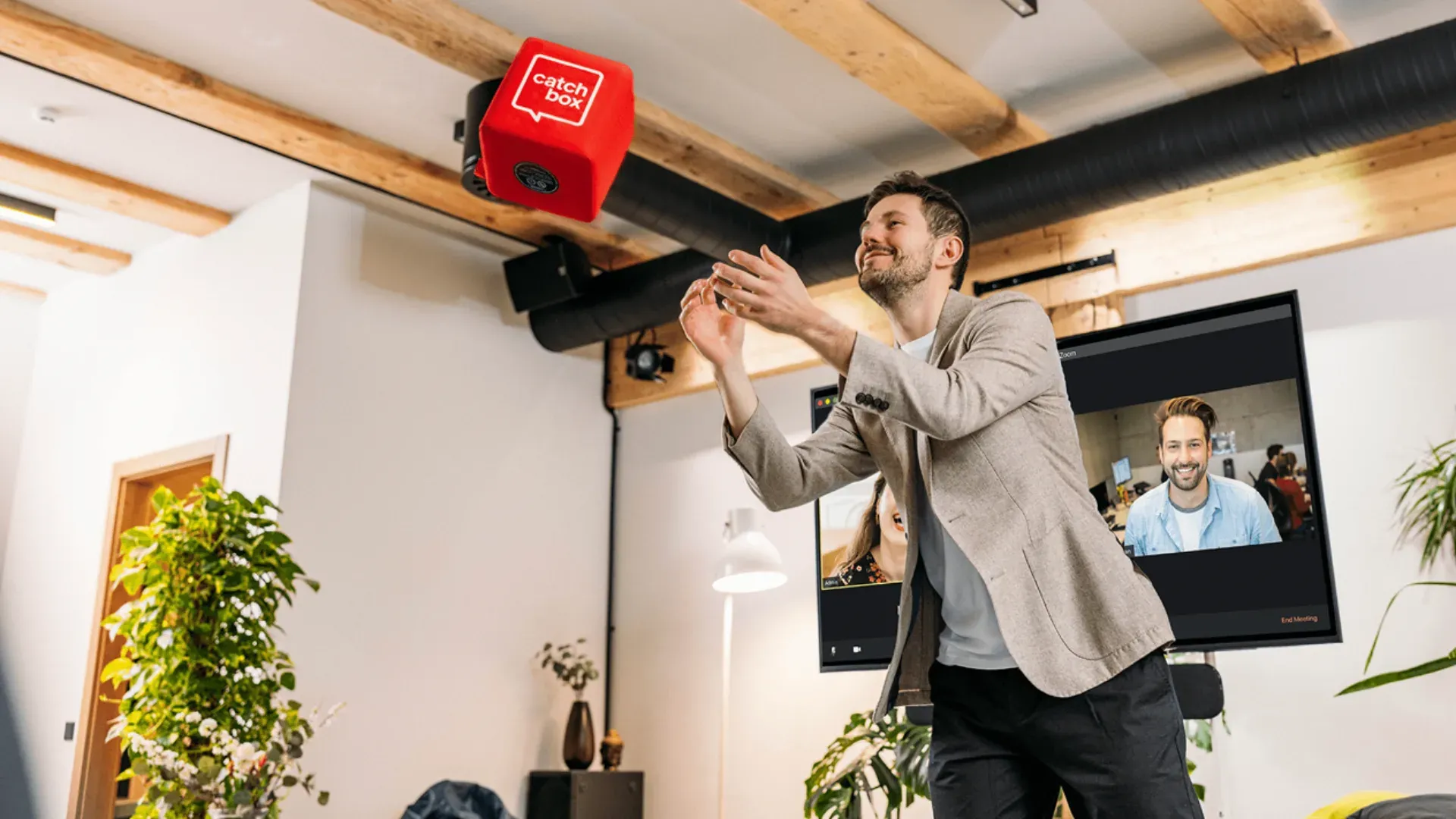 Homem a apanhar um cubo de microfone vermelho numa sala de reuniões, com participantes remotos num ecrã ao fundo.