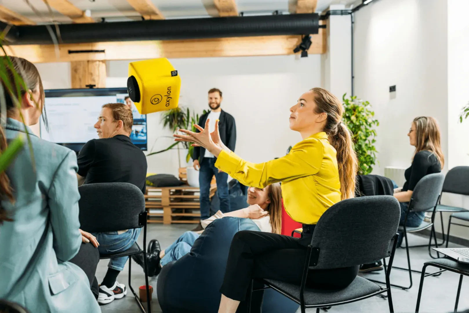 Sessão num espaço de trabalho, com as pessoas sentadas em roda, um cubo de microfone amarelo no ar e uma participante de braços estendidos para o apanhar.