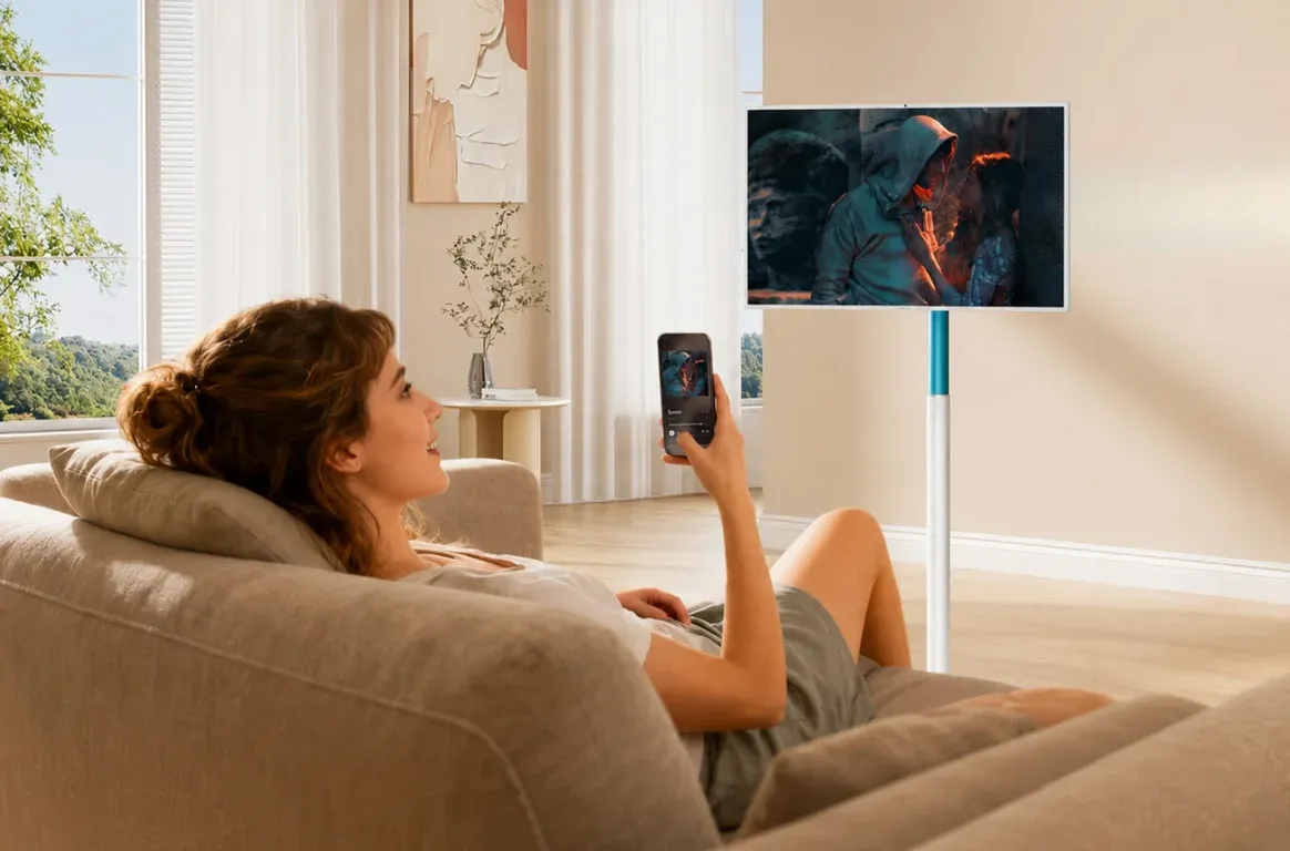Numa sala de estar, uma mulher no sofá segura o telemóvel, que mostra o mesmo vídeo que o ecrã Smart Rotate na horizontal, ao fundo.